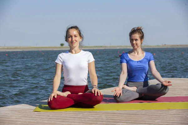 Zwei junge Frauen beim Yoga in der Natur. Fitness, Sport, Yoga und gesundes Lebensstil-Konzept - Gruppe von Yoga-Teilnehmern posiert bei Sonnenuntergang auf der Seebrücke — Stockfoto