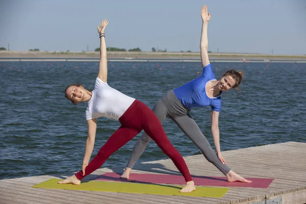 Zwei junge Frauen beim Yoga in der Natur. Fitness, Sport, Yoga und gesundes Lebensstil-Konzept - Gruppe von Yoga-Teilnehmern posiert bei Sonnenuntergang auf der Seebrücke — Stockfoto