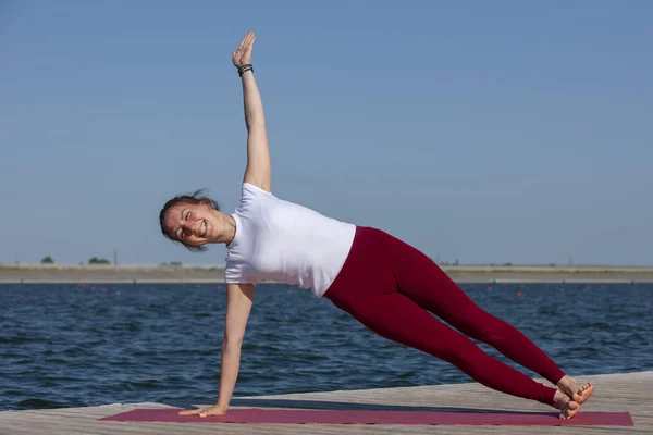 Chica practicando yoga cerca del lago, disfrutando de un buen día en la naturaleza y la energía positiva . — Foto de Stock