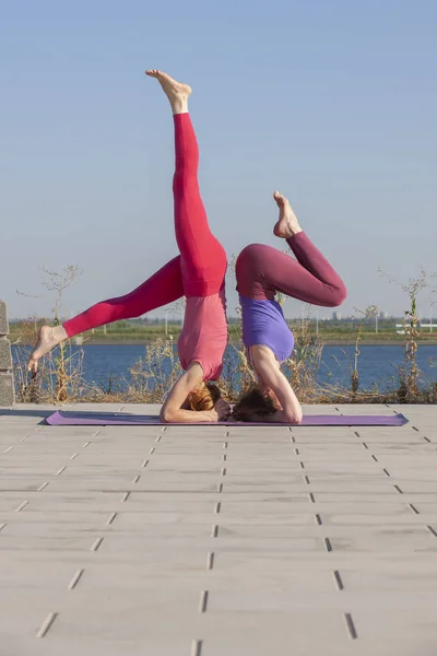 Zwei kaukasische Frauen in Strumpfhosen und Turnschuhen machen Yoga-Übungen auf dem Hintergrund des Flusses. — Stockfoto