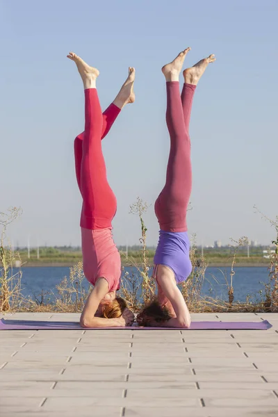 Due donne caucasiche in calzamaglia e scarpe da ginnastica che fanno esercizi di yoga sullo sfondo del fiume . — Foto Stock