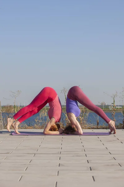 Zwei kaukasische Frauen in Strumpfhosen und Turnschuhen machen Yoga-Übungen auf dem Hintergrund des Flusses. — Stockfoto