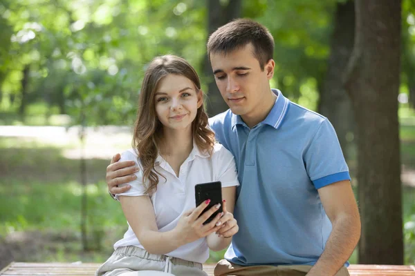 Пара обменивающихся СМИ в смартфоне, сидящих на скамейке в парке . — стоковое фото