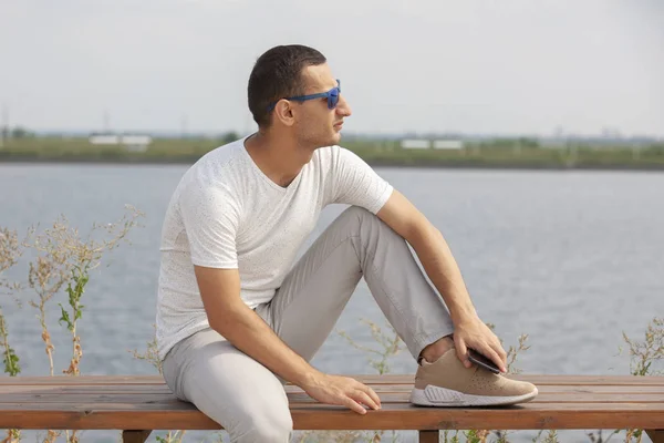 Happy young handsome man sitting on the bench outdoors and using smartphone — Stock Photo, Image