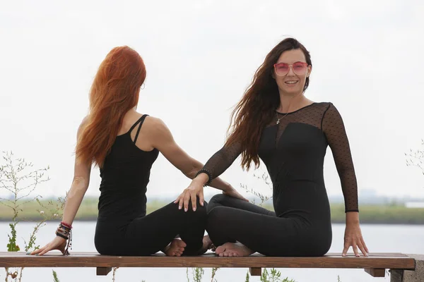 Dos mujeres jóvenes haciendo yoga en la naturaleza. Fitness, deporte, yoga y estilo de vida saludable - grupo de personas que hacen yoga posan en el muelle del lago al atardecer —  Fotos de Stock