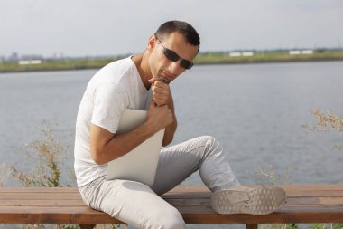 Bir dizüstü bilgisayar ile deniz kenarında oturan yakışıklı genç adam.