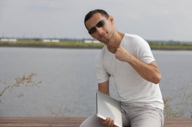 Bir dizüstü bilgisayar ile deniz kenarında oturan yakışıklı genç adam.