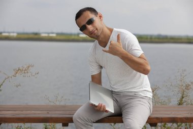 Bir dizüstü bilgisayar ile deniz kenarında oturan yakışıklı genç adam.