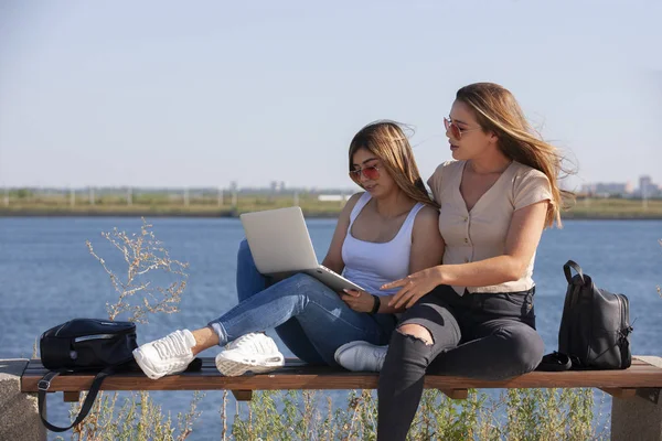 İki genç kız arkadaş bir bankta otururken bir dizüstü bilgisayar ekrana gösterirken diğeri dışarıda gülüyor. — Stok fotoğraf