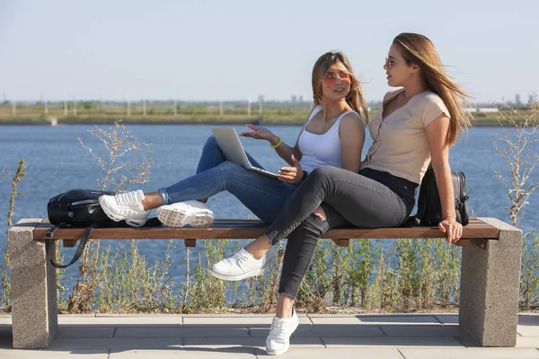 Två unga kvinnliga flickvänner ha kul medan du sitter på en bänk medan man visar att skärmen på en bärbar dator medan en annan är att skratta utanför. — Stockfoto