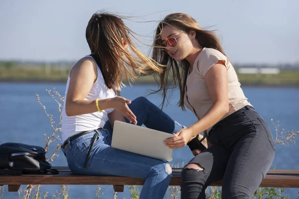 Två vackra unga kaukasiska flickor ser förvånad över att en bärbar dator när du sitter ute i parken på en bänk. — Stockfoto