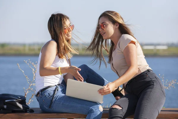 Två vackra unga kaukasiska flickor ser förvånad över att en bärbar dator när du sitter ute i parken på en bänk. — Stockfoto