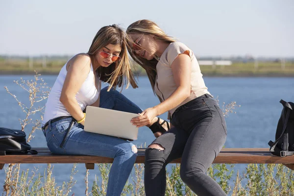 Två vackra unga kaukasiska flickor ser förvånad över att en bärbar dator när du sitter ute i parken på en bänk. — Stockfoto