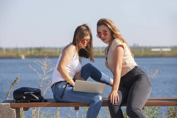 Två vackra unga kaukasiska flickor ser förvånad över att en bärbar dator när du sitter ute i parken på en bänk. — Stockfoto