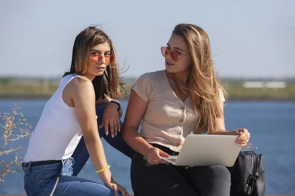 Två flickor sitta på en bänk i parken sommaren. De tittar på den bärbara skärmen. — Stockfoto