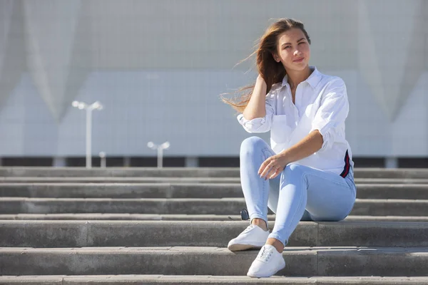 Beyaz moda spor ayakkabı kot pantolon beyaz bir t-shirt genç Amerikalı kadın merdivenlerde oturan rahatlatır. Sevimli Avrupa kız modeli geri kalanı sahiptir. Bahar tarzı kadın giyim.