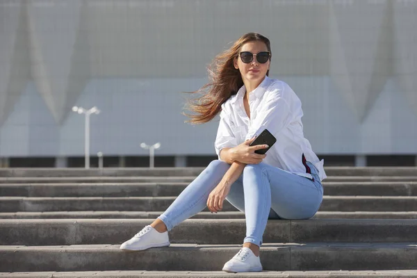 Jeune femme americaine en T shirt blanc en jeans en baskets blanches tendance se detend assis