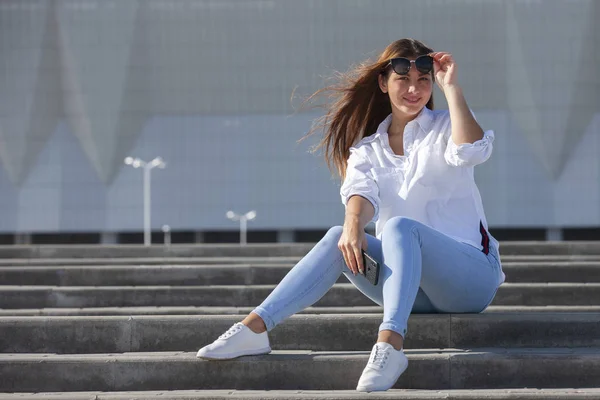 Молодая американка в белой футболке в джинсах в белых модных кроссовках расслабляется, сидя на ступеньках. Симпатичная европейская девушка-модель наслаждается остальным. Весенний стиль женской одежды . — стоковое фото