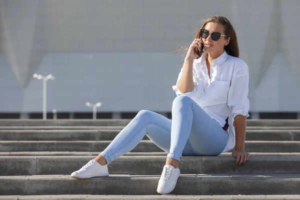 Ung amerikansk kvinna i en vit T-shirt i jeans i vita trendiga sneakers slappnar av sittande på trappan. Söt europeisk tjej modell åtnjuter resten. Vårstil damkläder. — Stockfoto