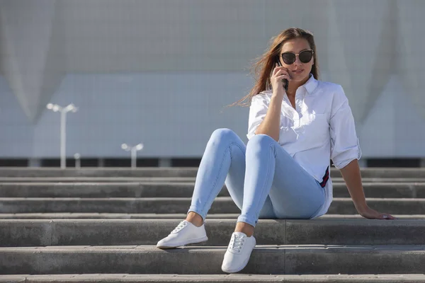 Молодая американка в белой футболке в джинсах в белых модных кроссовках расслабляется, сидя на ступеньках. Симпатичная европейская девушка-модель наслаждается остальным. Весенний стиль женской одежды . — стоковое фото
