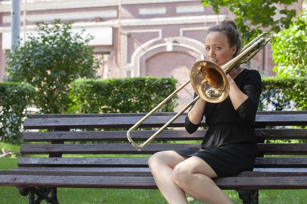 Dívka se učí hrát na trombon. Dívka hraje sedět na lavičce v parku. — Stock fotografie