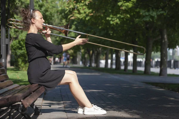 Chica aprendiendo a tocar el trombón. Chica juega sentado en un banco del parque . — Foto de Stock
