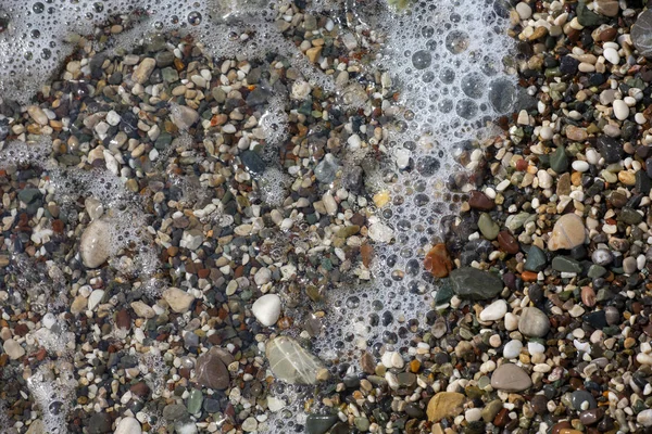 Espuma blanca delgada y las muchas burbujas pequeñas en el agua de mar brillante con los guijarros coloridos debajo —  Fotos de Stock