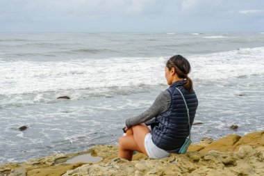 Genç Asyalı bir kadın uçurumda oturuyor ve uzaklara, denize bakıyor. Sahilde denize bakan güzel bir kız. İlham verici bir an. Ufuk çizgisine bakan genç bir kadının portresi