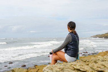 Genç Asyalı bir kadın uçurumda oturuyor ve uzaklara, denize bakıyor. Sahilde denize bakan güzel bir kız. İlham verici bir an. Ufuk çizgisine bakan genç bir kadının portresi