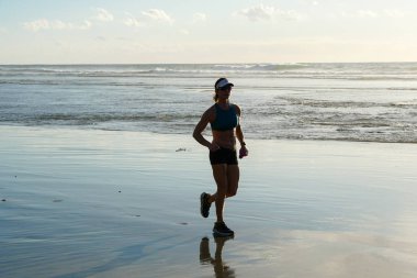 Gün batımında plajda koşu spor güzel genç kadın yan görünümü. Kadın koşucu sahilde açık havada koşu yapıyor. San Elijo Eyalet Plajı, Encinitas, San Diego, Kaliforniya. Abd