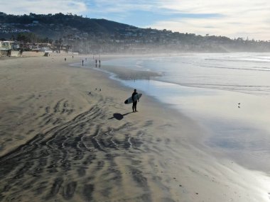 Sahilde sörf tahtası tutan ıslak takım elbiseli sörfçühavadan görünümü. Sörfçü su dışarı gidiyor ve gün batımı sırasında yürüyüş, La Jolla Beach, San Diego, California, Abd.