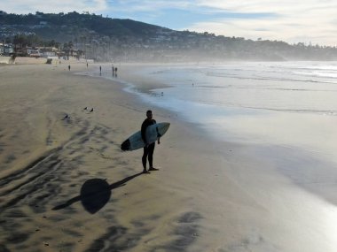 Sahilde sörf tahtası tutan ıslak takım elbiseli sörfçühavadan görünümü. Sörfçü su dışarı gidiyor ve gün batımı sırasında yürüyüş, La Jolla Beach, San Diego, California, Abd.