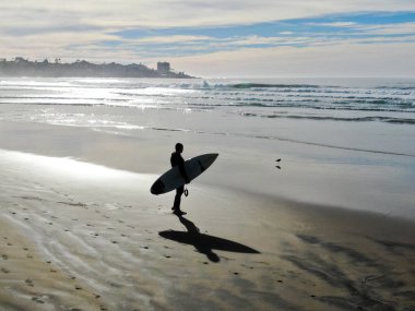 Sahilde sörf tahtası tutan ıslak takım elbiseli sörfçühavadan görünümü. Sörfçü su dışarı gidiyor ve gün batımı sırasında yürüyüş, La Jolla Beach, San Diego, California, Abd.