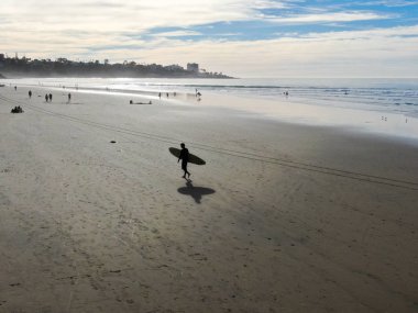 Sahilde sörf tahtası tutan ıslak takım elbiseli sörfçühavadan görünümü. Sörfçü su dışarı gidiyor ve gün batımı sırasında yürüyüş, La Jolla Beach, San Diego, California, Abd.