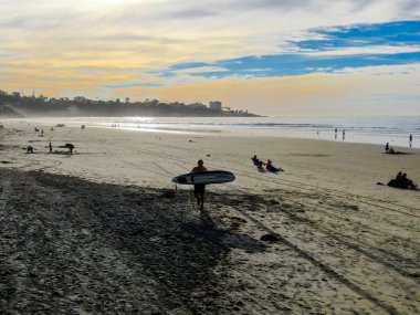 Sahilde sörf tahtası tutan ıslak takım elbiseli sörfçühavadan görünümü. Sörfçü su dışarı gidiyor ve gün batımı sırasında yürüyüş, La Jolla Beach, San Diego, California, Abd.
