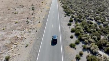 Tozlu, kuru çöl arazisinin ortasında asfalt yolda giden arabanın hava görüntüsü.