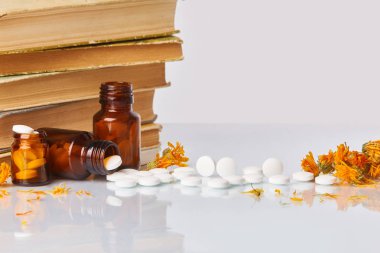 Tabletler ve hapları kadife çiçeği (Calendula) ve eski kitaplar beyaz ayna arka plan üzerinde beyaz. Sağlık kavramı. Homeopatik kavramı. Cam kahverengi şişe dağınık takviyeleri  