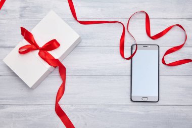 Kırmızı kurdeleli beyaz hediye kutusu ve siyah ahşap arka planda akıllı telefon.. 