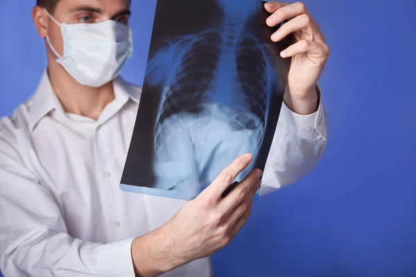 Male doctor in mask and  white coat holding  x-ray or roentgen of lungs, fluorography,  image  on blue background