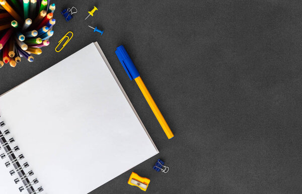 Открыть белый чистый спиральный блокнот с ручкой, цветные карандаши, точилка, сгустки бумаги, скрепки и нажать булавки на пустой темный вид сверху
