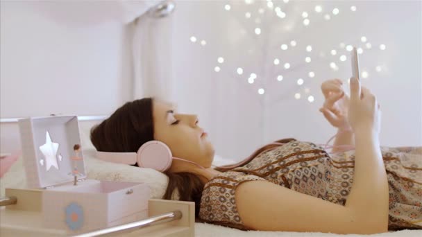 かなりCaucasian女の子リッスン音楽とともにヘッドフォンとスマートフォン上のベッド — ストック動画