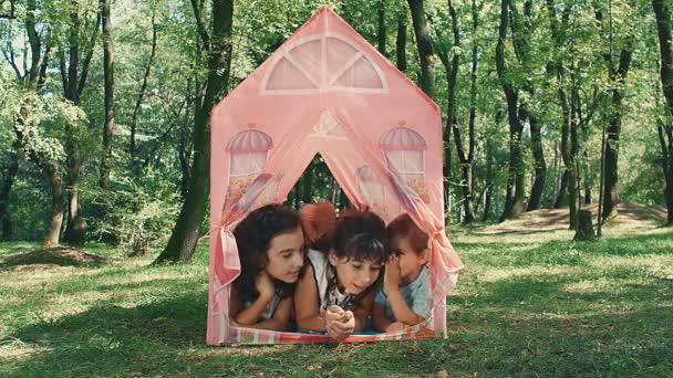 2人の女の子と男の子が公園でテントのおもちゃに横になって噂話をしています — ストック動画
