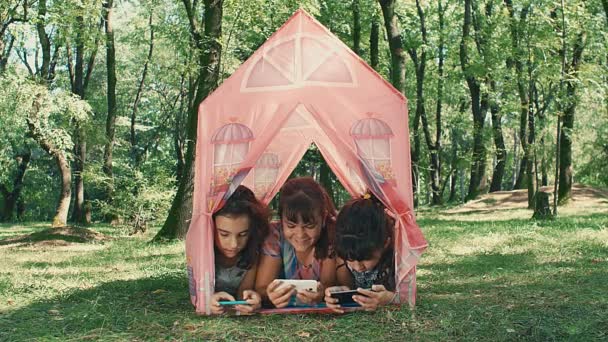 三人のガールフレンド遊びますスマートフォンで公園に横たわっているテントおもちゃ — ストック動画