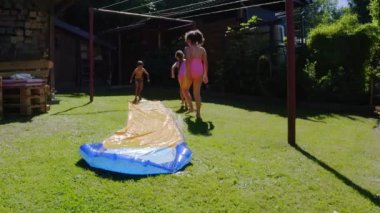 Küçük beyaz çocuklar bahçede su kaydırağında eğleniyorlar. Çocuklar ve çocuklar için serin yaz tatilleri. Suda oyunlar. 4k