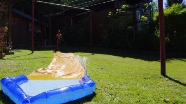 Küçük mutlu bir beyaz çocuk bahçede su kaydırağında eğleniyor. Çocuklar ve çocuklar için serin yaz tatilleri. Suda oyunlar. 4k