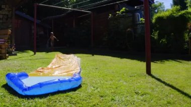 Küçük mutlu bir beyaz çocuk bahçede su kaydırağında eğleniyor. Çocuklar ve çocuklar için serin yaz tatilleri. Suda oyunlar. 4k