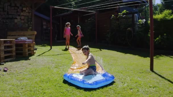 幸せな小さな白人の子供たちは庭のウォータースライドで楽しんでいます 子供や子供のための涼しい夏の休日 水の上のゲーム — ストック動画