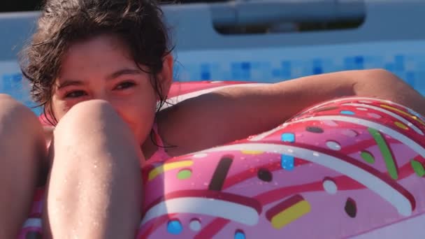12歳の幸せなかわいい白人の女の子は庭のスイミングプールで膨脹可能なドーナツサークルに横たわっています 子供や子供のための涼しい夏の休日 水の上のゲーム ピンクのドーナツ 4Kスローモーション — ストック動画