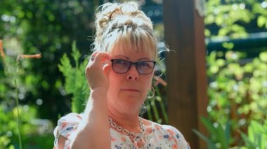 Gözlüklü, yeşil bahçe arka planındaki kameraya bakan 60 yaşındaki sarışın kadının portresi. 4k ağır çekim