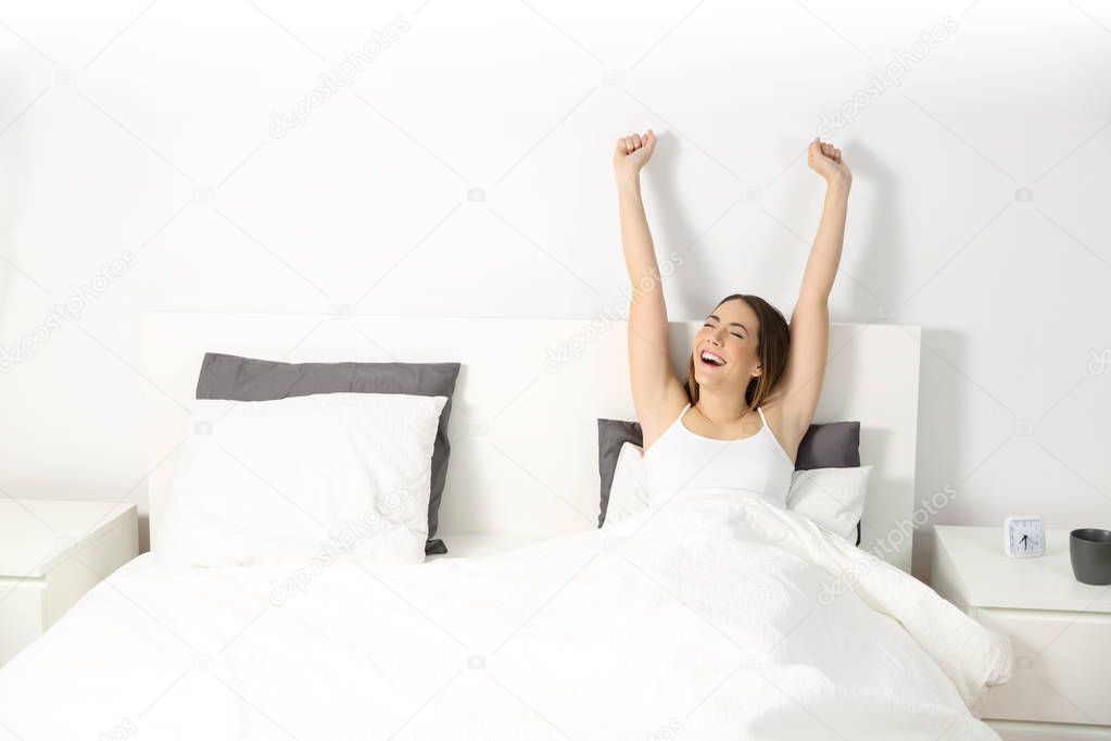 Excited woman sitting on a bed raising arms waking up in the morning with copy space